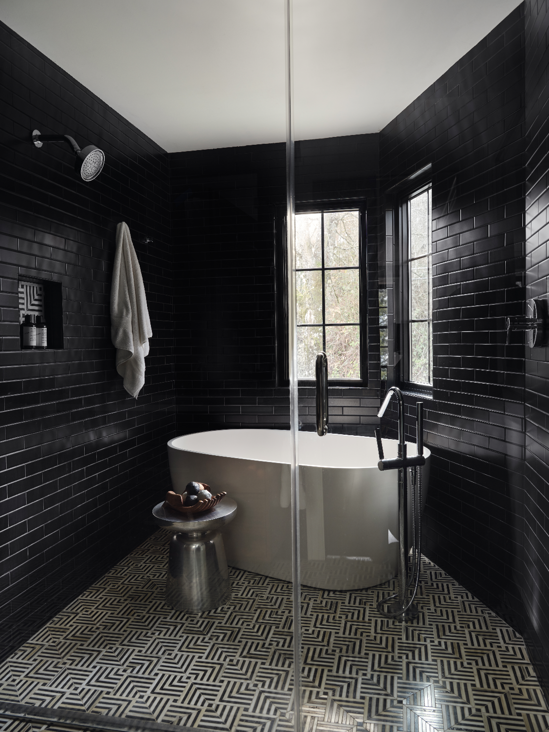 Luxury walk-in shower clad in glossy black tile with a freestanding soaking tub, geometric tile flooring, and black-framed windows.